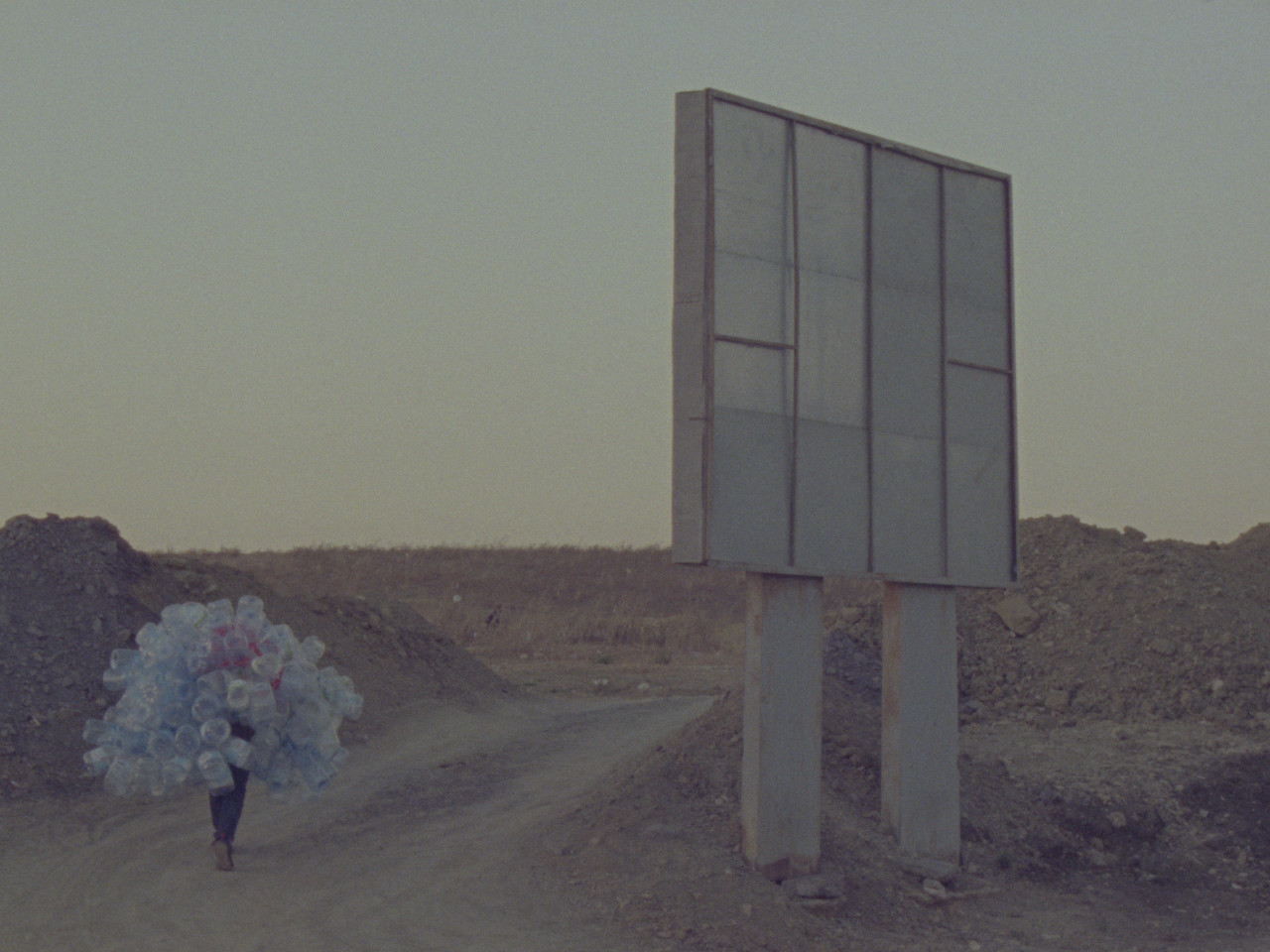 A color film still depicting a person who walks up a dirt path carrying a myriad of empty plastic bottles. To the person's right is a billboard facing away from the camera