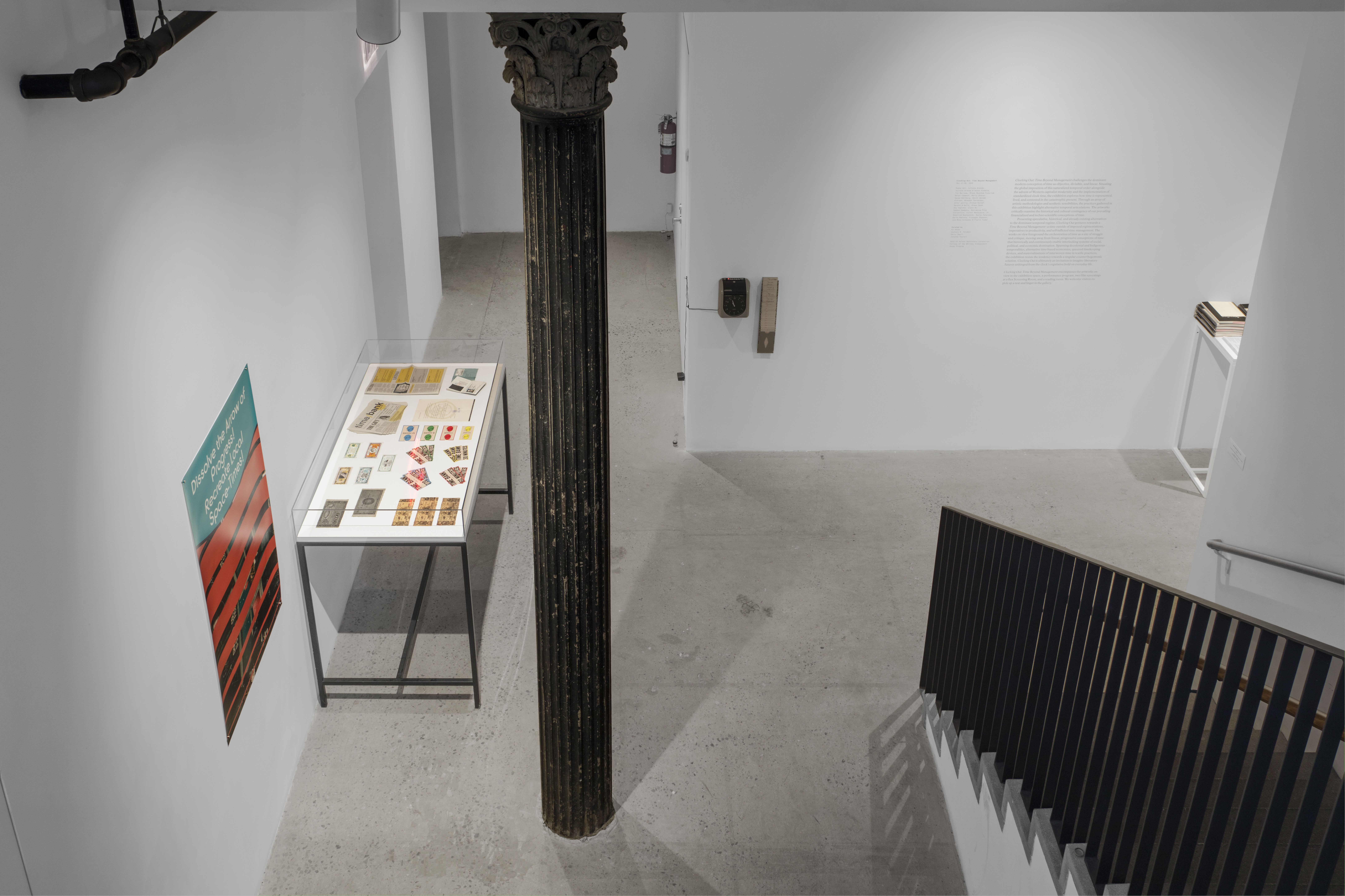 Photograph of a well-lit gallery space at the bottom of a staircase, with a glass vitrine on the left wall containing ephemera.