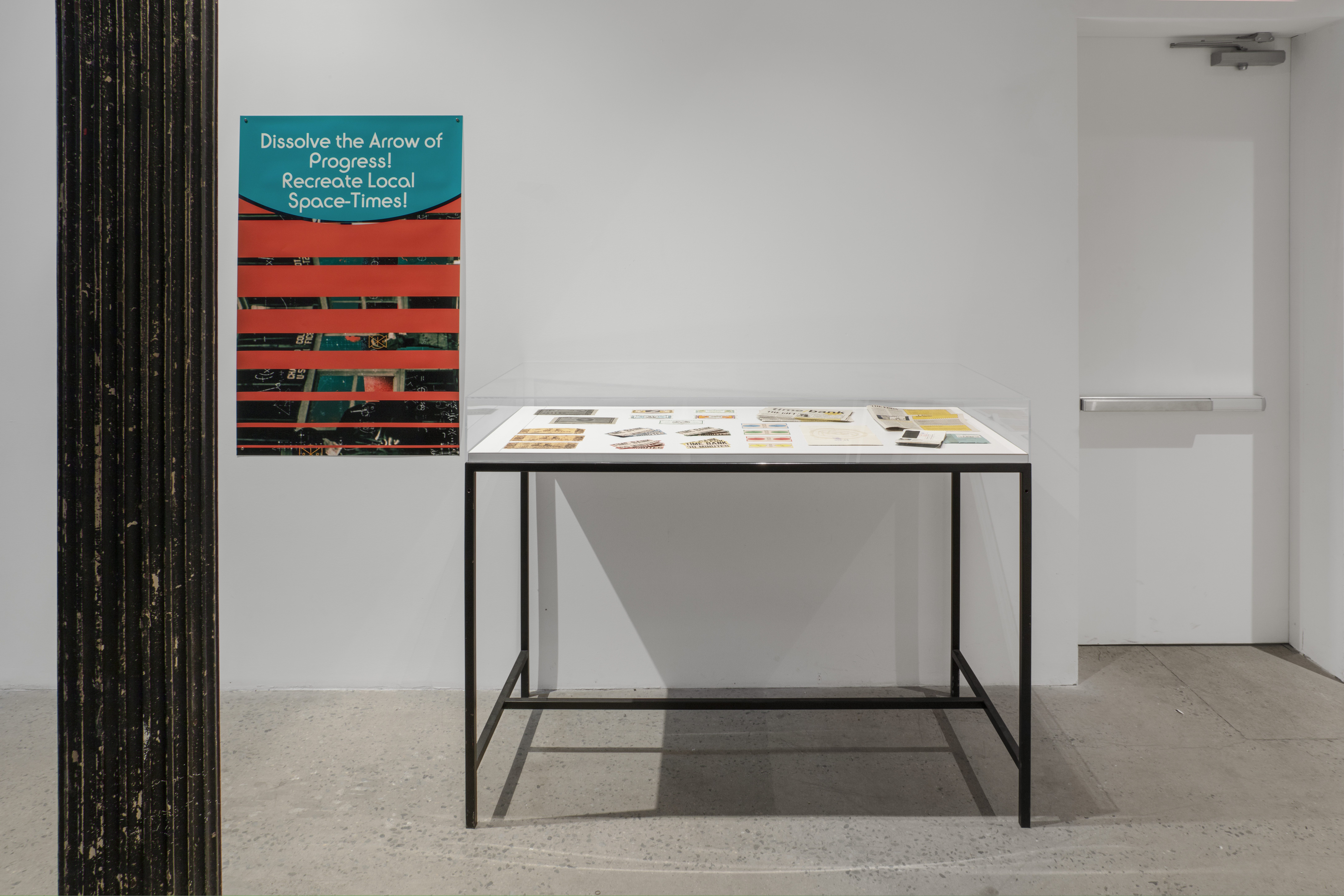 Photograph of a glass vitrine sitting against a wall, containing ephemera, and a graphic poster on the left that reads "Dissolve the Arrow of Progress! Recreate Local Space-Times!"