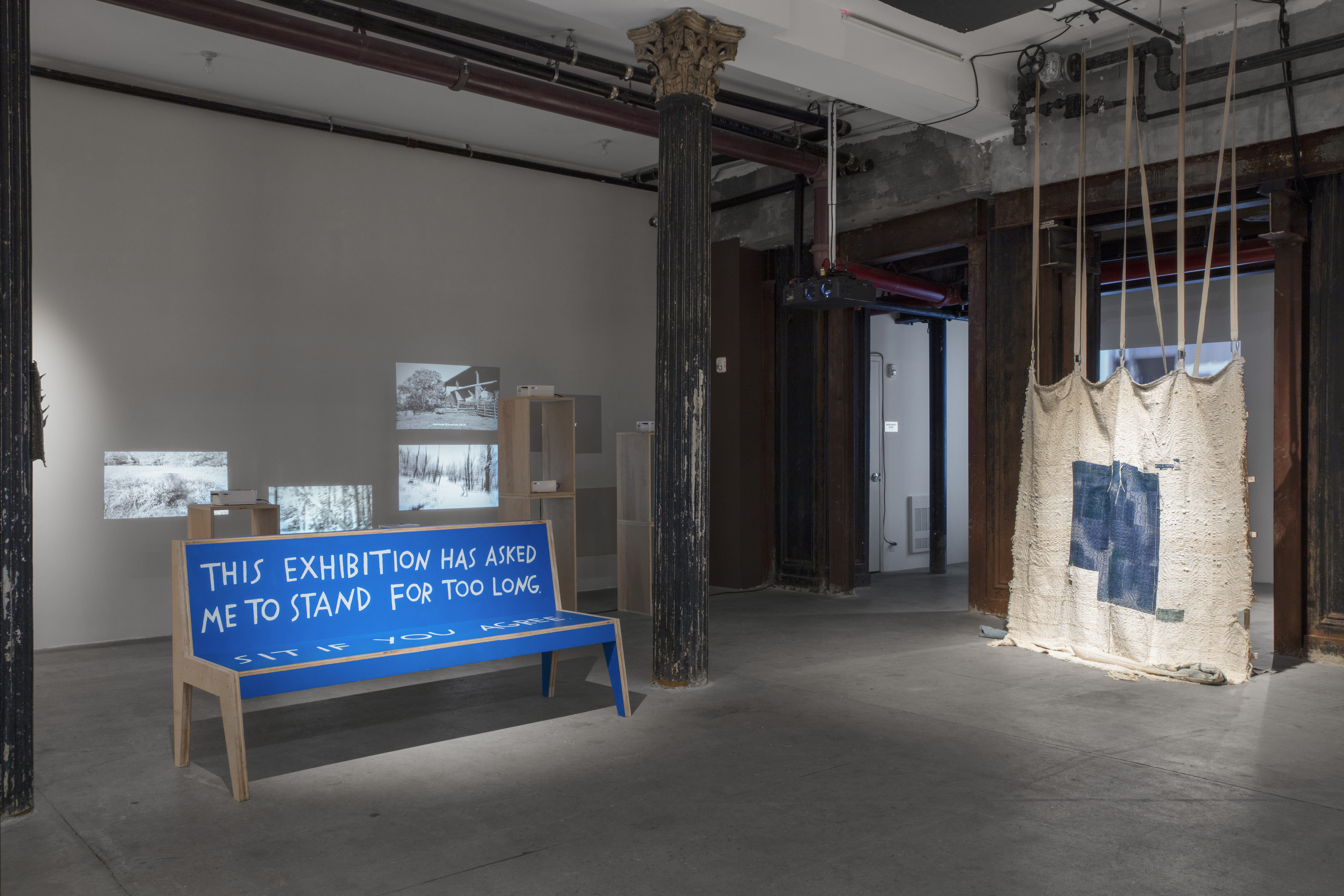 Photograph of a corner of a basement gallery. A wooden bench, painted blue on the surface, sits between two columns. The back of the bench reads "THIS EXHIBITION HAS ASKED / ME TO STAND FOR TOO LONG." A woven piece hangs from the ceiling to the right. Behind the bench are projectors facing the wall at varying heights.