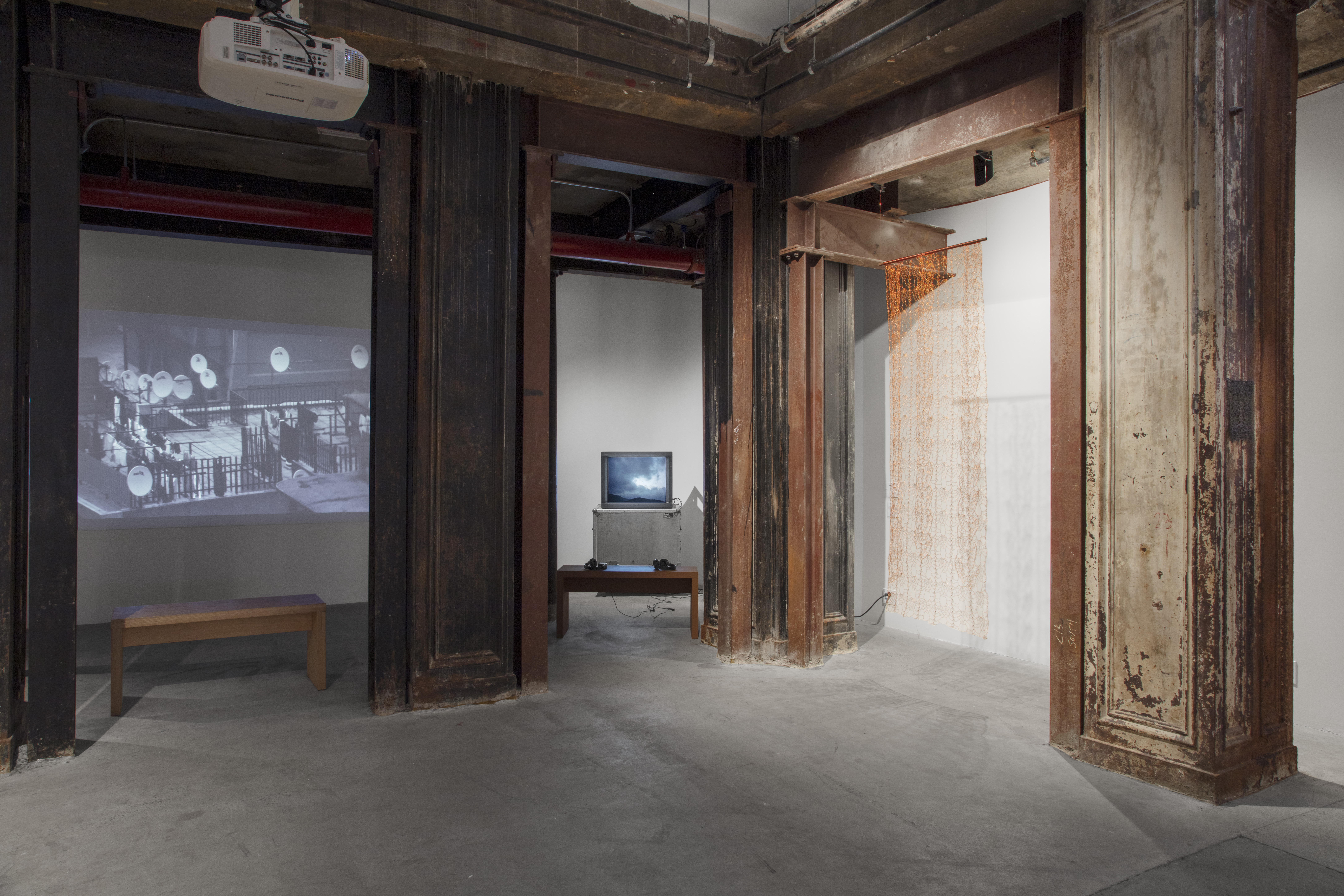 Photograph of a corner of a basement gallery space with large columns along the back. Behind the columns are benches placed in front of video displays. On the right is a CRT TV monitor, and to the left is a projected video.