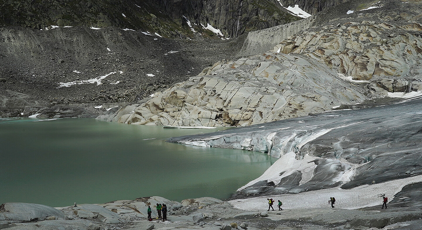 Film still depicting seven hikers near the bottom. A mountainous landscape occupies most of the screen with a grey-green lake on the left.